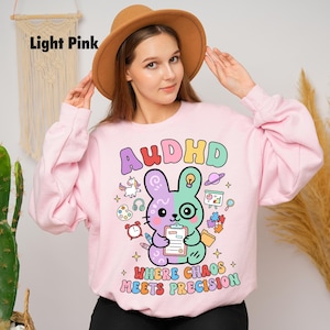 May include: Pale pink sweatshirt featuring the word "AUDHD" in colourful letters and a cartoon bunny holding a clipboard. The bunny is half purple and half green. The text "WHERE CHAOS MEETS PRECISION" is below the bunny.