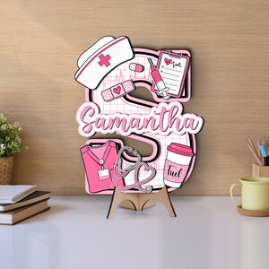 Customizable wooden door sign with two layers, showcasing a large letter S with medical motifs in pink. Includes nurse hat, clipboard, syringe, and personalized name Samantha. Ideal for adding a personal touch to a nurse's workspace.