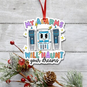 May include: A white Christmas ornament with a cartoon illustration of a medical infusion pump with the text "MY ALARMS WILL HAUNT your dreams". The pump has a blue screen with a cartoon face and the text "FAST FLOW FOR HUMAN USE ONLY".