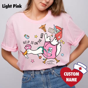 May include: Pale pink t-shirt with a cartoon duck in a nurse's uniform, including a stethoscope and medical equipment. The phrase "HELP IS ON THE WAY" is printed on the shirt. A heart-shaped graphic with a nurse's hat and "CUSTOM NAME" is also visible.