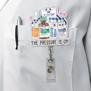 May include: A badge reel featuring cartoon illustrations of medical vials and a bag of dextrose. The text "THE PRESSURE IS ON" is displayed below the illustrations. The reel is attached to a white pocket.