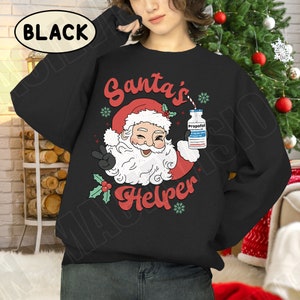Puede incluir: Sudadera negra con un gr&aacute;fico de Pap&aacute; Noel sosteniendo una botella etiquetada "Propofol" y el texto "Santa's Helper". Pap&aacute; Noel lleva un gorro rojo y hace la se&ntilde;al de la paz. La palabra "BLACK" est&aacute; en un &oacute;valo blanco.