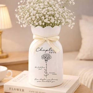 May include: A white ceramic vase with a scalloped rim, tied with a cream-colored ribbon, and filled with white flowers. The vase has black text that reads "Chapter Fifty" and "Olivia", with a flower illustration and other text.