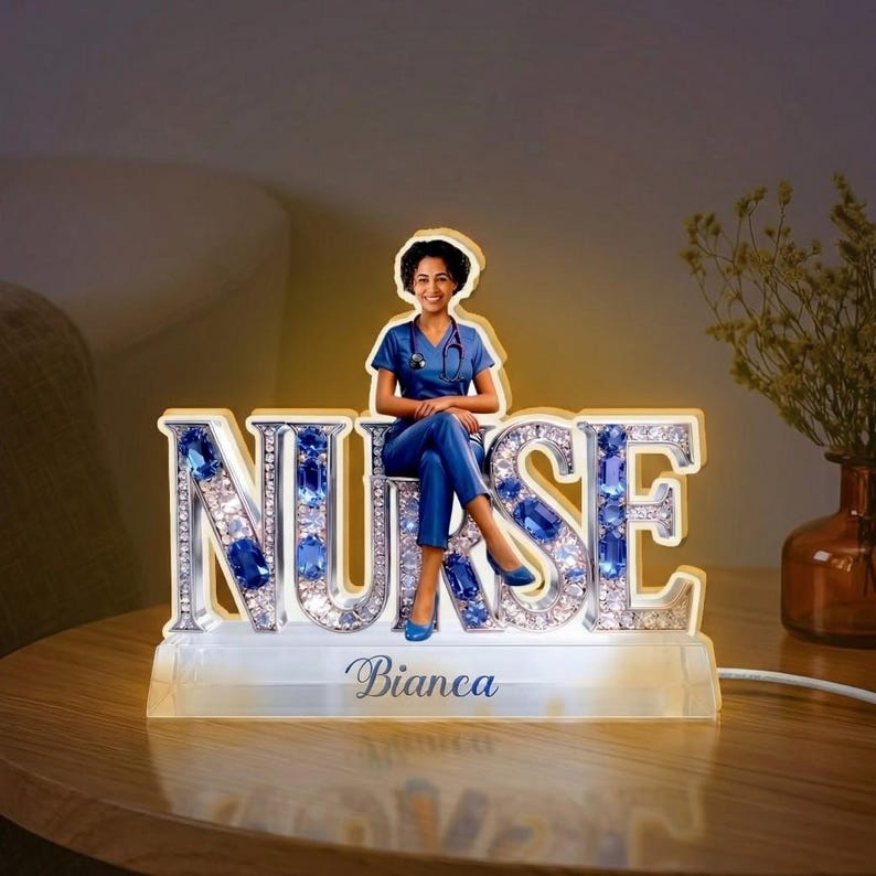 May include: A decorative illuminated sign with the word "NURSE" spelled out in large, sparkling letters. The letters are embellished with blue gems. A figure of a nurse in blue scrubs sits atop the letters. The name "Bianca" is written below the letters.