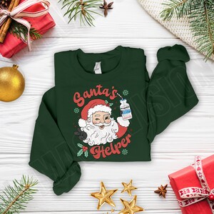 Puede incluir: Sudadera verde oscuro con un gr&aacute;fico de Pap&aacute; Noel sosteniendo un vial etiquetado "Propofol" y el texto "Santa's Helper". El dise&ntilde;o incluye un gorro de Pap&aacute; Noel rojo, acebo y copos de nieve. Decoraciones navide&ntilde;as.