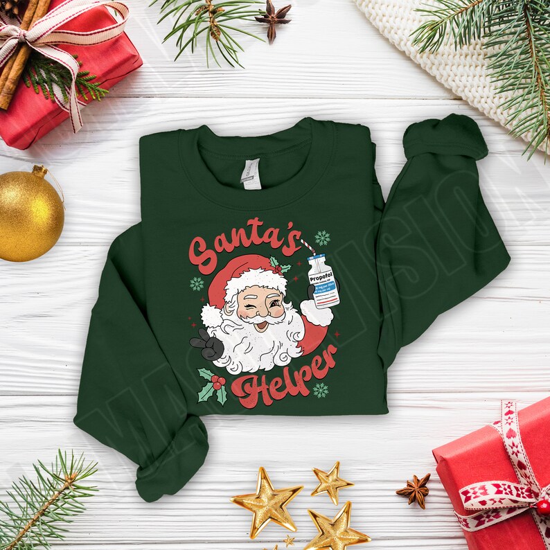 Puede incluir: Sudadera verde oscuro con un gr&aacute;fico de Pap&aacute; Noel sosteniendo un vial etiquetado "Propofol" y el texto "Santa's Helper". El dise&ntilde;o incluye un gorro de Pap&aacute; Noel rojo, acebo y copos de nieve. Decoraciones navide&ntilde;as.