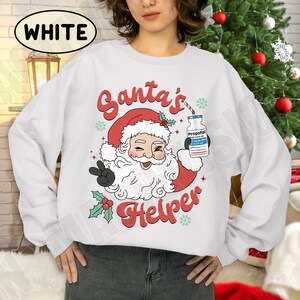 Puede incluir: Sudadera blanca con un dise&ntilde;o de Pap&aacute; Noel gui&ntilde;ando un ojo y haciendo la se&ntilde;al de la paz. Pap&aacute; Noel lleva un gorro rojo y sostiene un frasco con la etiqueta "Propofol". El texto "Santa's Helper" est&aacute; en rojo.