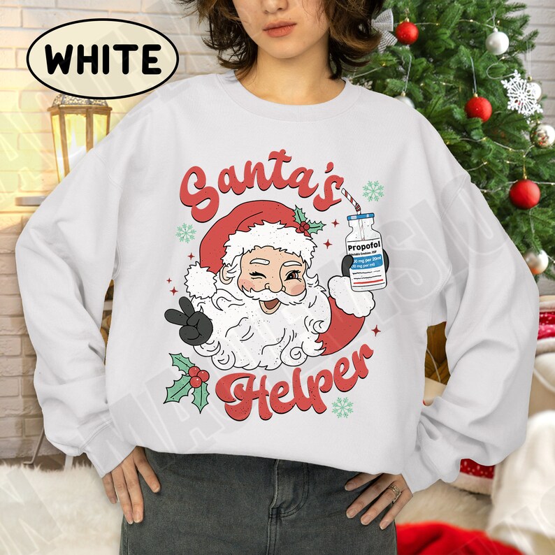 Puede incluir: Sudadera blanca con un dise&ntilde;o de Pap&aacute; Noel gui&ntilde;ando un ojo y haciendo la se&ntilde;al de la paz. Pap&aacute; Noel lleva un gorro rojo y sostiene un frasco con la etiqueta "Propofol". El texto "Santa's Helper" est&aacute; en rojo.