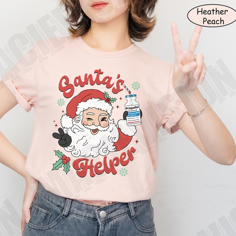 Puede incluir: Camiseta color melocot&oacute;n con un gr&aacute;fico de Pap&aacute; Noel gui&ntilde;ando un ojo y haciendo el signo de la paz. El texto dice "Santa's Helper" en rojo, con una botella de Propofol. La camiseta tiene mangas enrolladas.