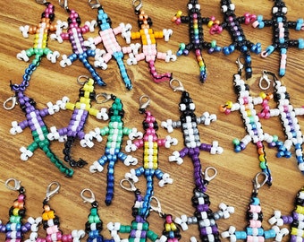 Può includere: Una collezione di 15 portachiavi a forma di lucertola con perline colorate. Ogni portachiavi è realizzato con piccole perline di plastica di vari colori, tra cui blu, verde, viola, rosa, giallo, arancione, nero e bianco. Le lucertole sono tutte in una posa simile, con le zampe distese e le code arrotolate.