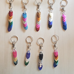 Op de afbeelding: Tien sleutelhangers met verschillende gekleurde hartvormige bedels. Elke sleutelhanger heeft een zilveren ring en een zilveren ketting. De bedels zijn in een rij gerangschikt, waarbij de kleuren van de bedels afwisselen.