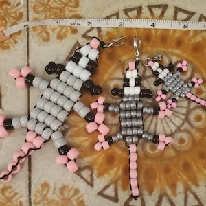 May include: Three beaded lizard keychains, each with a different color scheme. The first is gray and white with pink accents, the second is gray with pink accents, and the third is gray with pink accents. The keychains are made of small, round beads and have a silver loop for attaching to keys.