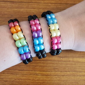 Può includere: Tre braccialetti colorati con perline e filo elastico nero. I braccialetti sono realizzati con perline arcobaleno e sono impilati sul polso di una persona.