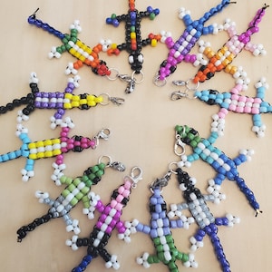 Op de afbeelding: Een verzameling van 10 kleurrijke kralen-hagedis sleutelhangers. Elke hagedis is gemaakt met verschillende gekleurde kralen en heeft een zwarte kralenstaart. De hagedissen zijn in een cirkel op een houten oppervlak gerangschikt.