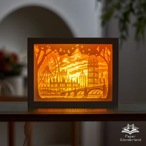 Könnte beinhalten: Ein beleuchteter Papier-Schnitt-Rahmen mit der Silhouette von London, einschließlich London Eye, Big Ben und Brücken. Das Kunstwerk wird von warmem orangefarbenem Licht beleuchtet, wodurch ein geschichteter Effekt entsteht. Der Text "Paper Wonderland" ist sichtbar.