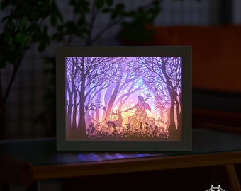 May include: A framed paper art scene of a forest with a cellist, deer, and trees, illuminated from within. The artwork uses layered paper to create depth, with a colour gradient from purple to orange. The frame is a neutral grey colour.