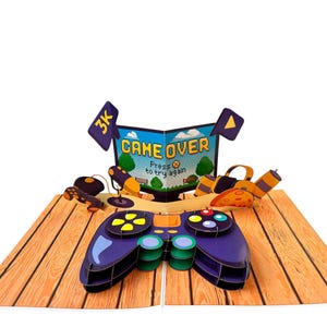 Puede incluir: Una tarjeta desplegable con temática de videojuegos. La tarjeta presenta un gran mando de juego morado y negro, una pantalla que muestra "GAME OVER" y otros accesorios de juego. La tarjeta está sobre una superficie de madera.