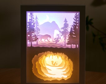 Könnte beinhalten: Eine weiße gerahmte Papierkunst-Leuchtlandschaft mit einem 3D-Wald, Mond und Tieren. Die Szene wird mit einem warmen gelben Licht beleuchtet, das einen magischen Schein erzeugt. Der untere Teil des Rahmens zeigt eine 3D-Papierblume mit einem Licht, das durch sie scheint.