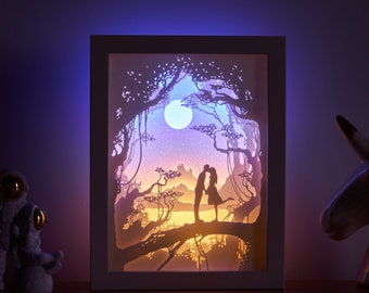 May include: A white frame containing a paper cut-out design of a couple kissing under a full moon in a forest. The design is illuminated from behind with a warm white light.
