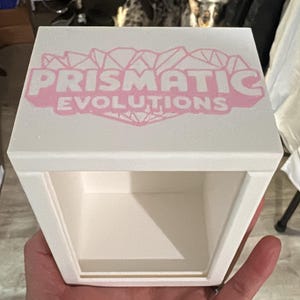May include: A white, open-front box with the text "PRISMATIC EVOLUTIONS" in pink across the top. The box is held in a person's hand. A dog is visible in the background.