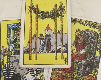 May include: Three tarot cards, "The Chariot", "Queen of Pentacles", and "Four of Wands". The cards are on a white surface. The Chariot card features a black and white sphinx, the Queen of Pentacles card features a woman sitting on a throne, and the Four of Wands card features a yellow background with three wands and a castle.