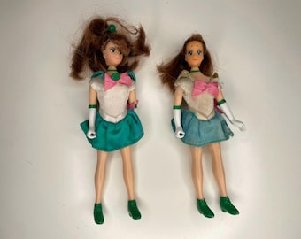 Puede incluir: Dos muñecas vintage con cabello castaño, que visten tops blancos, faldas turquesas y botas verdes. Cada muñeca tiene un lazo rosa y guantes blancos. Una muñeca tiene una cola de caballo y la otra tiene el pelo suelto.