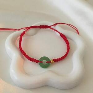 May include: A red string bracelet with a green circular jade pendant and a small gold bead. The bracelet is displayed on a white, flower-shaped dish. The jade is a light green color.