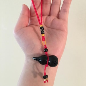 May include: A black gourd charm with a red string and black beads. The charm is tied to a red string with a gold and black tassel.