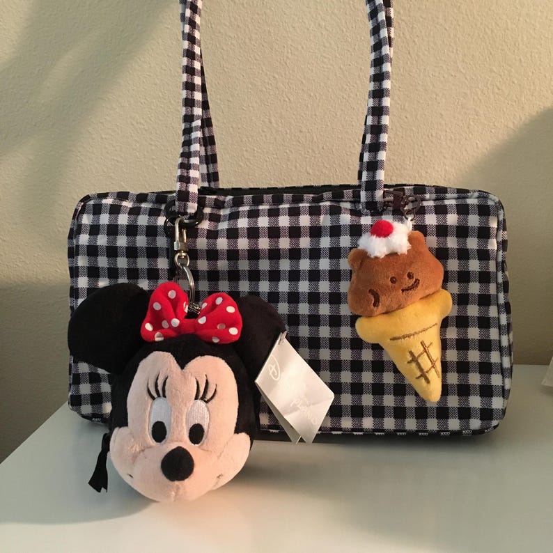 Pu&ograve; includere: Borsa a mano a quadretti bianchi e neri con manici coordinati. La borsa presenta un portachiavi in ​​peluche di Minnie Mouse con un fiocco a pois rossi e un portachiavi a forma di cono gelato orso marrone.