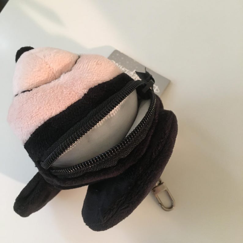 Pu&ograve; includere: Un portamonete a forma di panda in peluche, con cerniera nera e chiusura color argento. Il panda ha la testa rosa e nera, orecchie nere e corpo nero. Il portamonete &egrave; aperto, rivelando un interno bianco.