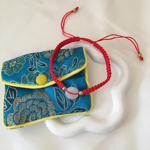 May include: A red braided bracelet with a light-colored stone and gold accents. The bracelet is next to a small blue pouch with a floral pattern and yellow trim. The pouch has a yellow button closure. The bracelet is on a white surface.