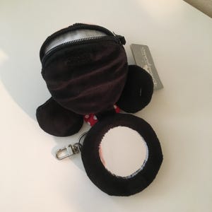 Pu&ograve; includere: Un portamonete in peluche nero con chiusura a cerniera, a forma di personaggio dei cartoni animati. Presenta un fiocco a pois rossi e bianchi e uno specchio rotondo attaccato con un fermaglio argentato. Un'etichetta Disneyland &egrave; visibile.