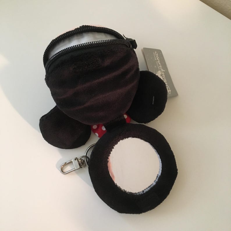 Pu&ograve; includere: Un portamonete in peluche nero con chiusura a cerniera, a forma di personaggio dei cartoni animati. Presenta un fiocco a pois rossi e bianchi e uno specchio rotondo attaccato con un fermaglio argentato. Un'etichetta Disneyland &egrave; visibile.