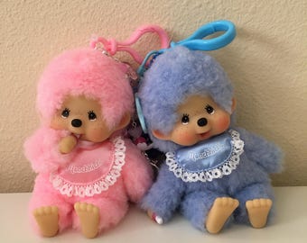 Könnte beinhalten: Zwei Monchhichi-Plüschpuppen, eine rosa und eine blaue, jeweils mit einem Lätzchen und einem Schnuller. Die rosa Puppe hat einen rosa Clip, und die blaue Puppe hat einen blauen Clip. Beide Puppen haben braune Augen und Sommersprossen.