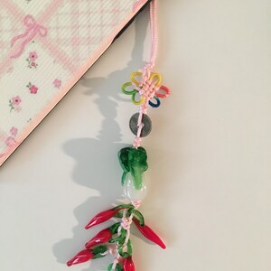 May include: A decorative charm with a pink cord, featuring a colorful flower, a coin, a green cabbage, and red chili peppers. The charm is hanging next to a pink and white floral patterned object.