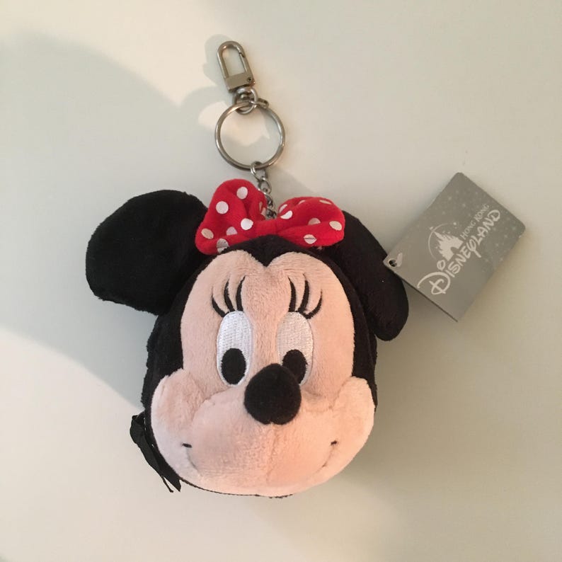 Pu&ograve; includere: Un portachiavi di peluche di Minnie Mouse. Il portachiavi presenta una testa nera con grandi orecchie, un viso rosa e un fiocco rosso a pois bianchi. Sono attaccati un moschettone e un anello in metallo argentato. &Egrave; attaccata anche un'etichetta grigia di Disneyland Hong Kong.