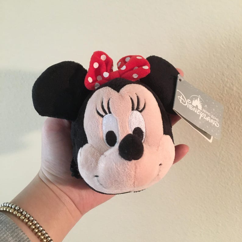 Pu&ograve; includere: Una testa di Minnie Mouse in peluche con orecchie nere, un fiocco rosso a pois bianchi e un viso rosa chiaro. L'etichetta riporta "Disneyland Hong Kong".