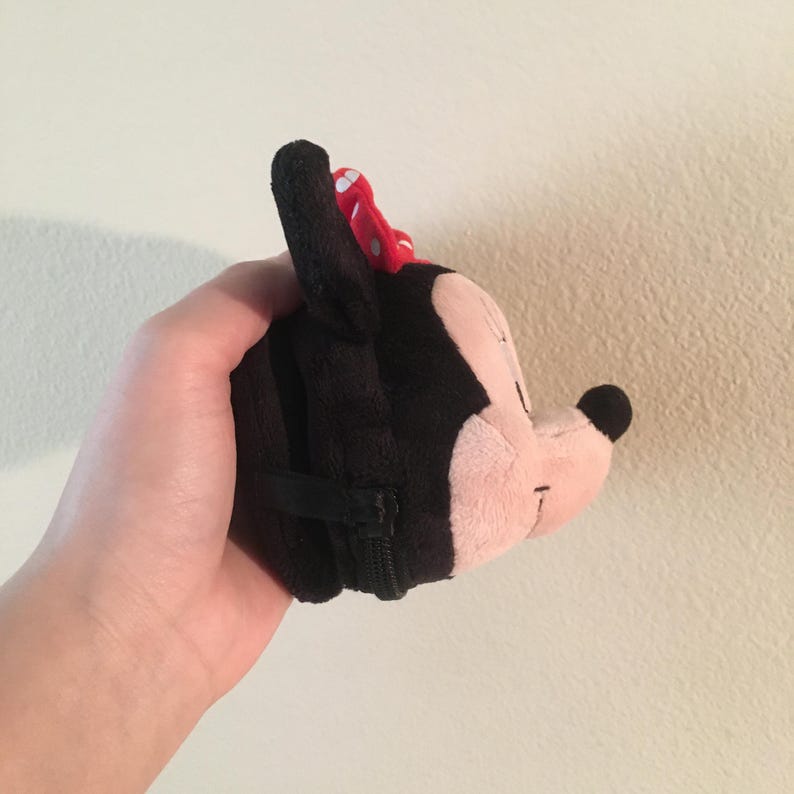 Pu&ograve; includere: Un portamonete a forma di Minnie Mouse in peluche nero con un fiocco rosso a pois. La borsa ha un viso rosa, orecchie nere e una chiusura con cerniera nera. La borsa &egrave; tenuta in una mano.