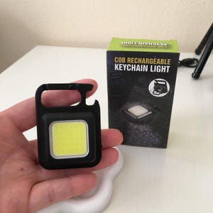 May include: A black COB rechargeable keychain light with a bright yellow square light. The light is held in a hand, next to its packaging, which reads "COB RECHARGEABLE KEYCHAIN LIGHT".