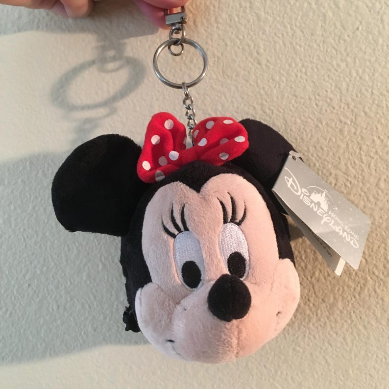Pu&ograve; includere: Un portachiavi in ​​peluche di Minnie Mouse con un fiocco rosso a pois bianchi. Il portachiavi ha un anello e una catena in metallo argentato. Il peluche ha orecchie nere e un viso rosa. &Egrave; attaccata un'etichetta Disney Hong Kong.