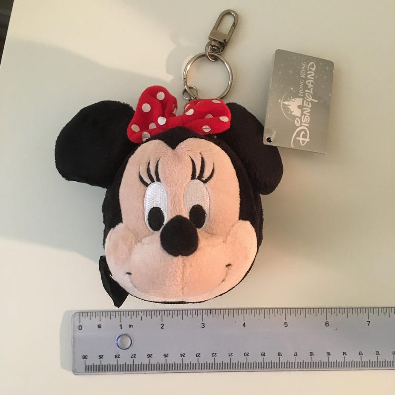 Pu&ograve; includere: Un portachiavi di Minnie Mouse in peluche. Il portachiavi presenta una testa nera con grandi orecchie nere, un fiocco rosso a pois bianchi e un viso rosa chiaro. Sono attaccati un fermaglio in metallo e un'etichetta Disneyland Hong Kong.