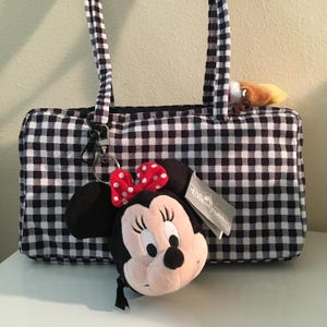 Pu&ograve; includere: Una borsa tote a quadretti bianchi e neri con manici neri. Un portachiavi di peluche di Minnie Mouse con un fiocco rosso e un'etichetta Disneyland &egrave; attaccato. Un piccolo peluche &egrave; visibile all'interno della borsa.