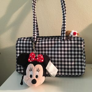 Pu&ograve; includere: Una borsa a quadretti bianchi e neri con manici abbinati. Un portachiavi di peluche di Minnie Mouse con un fiocco rosso a pois &egrave; appeso alla borsa. Un piccolo peluche &egrave; visibile all'interno della borsa.