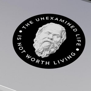 May include: Black and white sticker with a quote from Socrates: "The unexamined life is not worth living". The quote is surrounded by a circle with a bust of Socrates in the center.