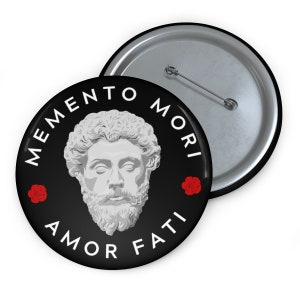 Pode incluir: Um botão preto com uma imagem branca do rosto de um homem e o texto "MEMENTO MORI AMOR FATI" ao redor da imagem. Há duas rosas vermelhas de cada lado da imagem.