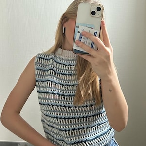 Op de afbeelding: Een wit en blauw gestreept, gehaakt, mouwloos topje met een hoge hals.