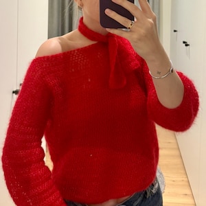 Op de afbeelding: Een rode, pluche gebreide trui met een strik om de hals. De trui heeft een licht off-shoulder ontwerp.