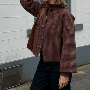 Puede incluir: Una chaqueta de color marrón oscuro de manga larga con un patrón de tejido de punto gofrado. La chaqueta presenta un cuello alto y un cierre frontal con cuatro botones de palanca de madera clara, sobre unos vaqueros azul oscuro.