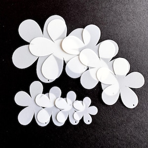 May include: White paper flower cutouts in various sizes, arranged on a black surface. The flowers have layered petals, creating a 3D effect. Each flower has a small hole, possibly for stringing or attaching. Ideal for crafting or scrapbooking.