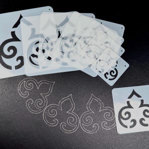 Peut inclure: Ensemble de pochoirs en plastique blanc avec des motifs décoratifs en volutes. Plusieurs pochoirs sont empilés et certains sont dispersés, avec le motif tracé sur la surface. Les pochoirs sont carrés et rectangulaires, sur fond sombre.